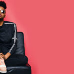 black woman sitting on a sofa wearing sunglasses and sportswear in a pink background